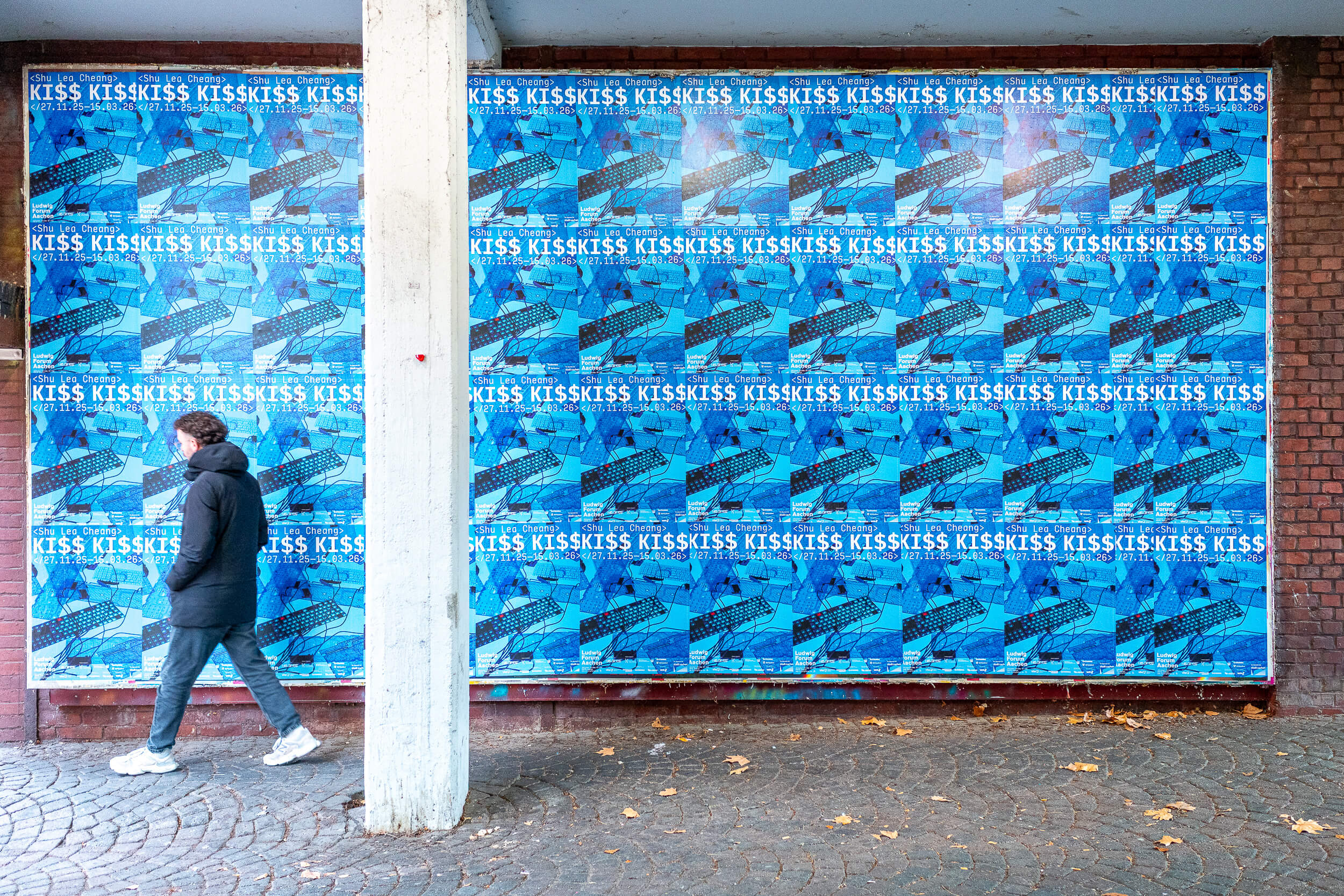 Büro Gestalten: Kommunikation zur Ausstellung »Shu Lea Cheang. KI$$ KI$$« (Ludwig Forum Aachen 2025/26), Poster im Rapport (mit vorbei gehender Person)